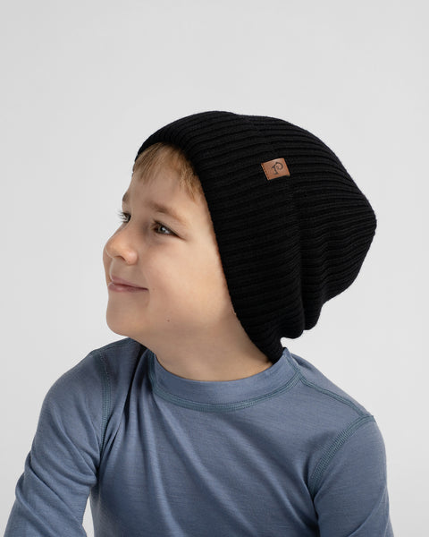 A child wearing a black ribbed slouchy beanie with a small brown brand patch, photographed against a light gray studio background.