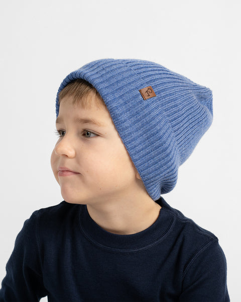 A young child wearing a ribbed knit blue beanie hat with a small brown logo patch, photographed against a clean light gray background.