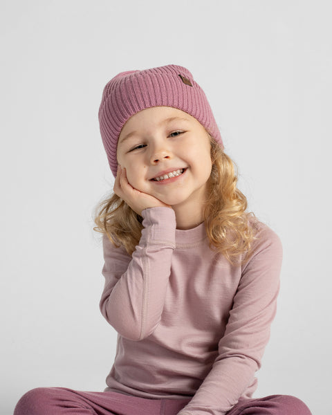 A young child wearing a ribbed knit beanie, a dusty pink long-sleeve top, and matching pink pants, sitting against a light gray studio background.