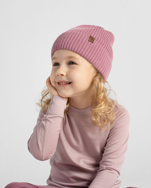 A young girl with blonde curly hair wearing a pink ribbed knit beanie and a matching dusty pink long-sleeve top, posing against a light gray studio background.