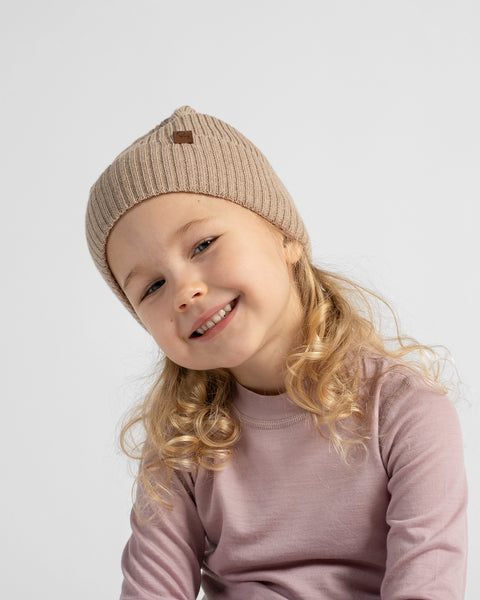 A young girl with blonde curly hair wearing a beige ribbed knit beanie with a small brown logo patch and a dusty pink long-sleeve shirt, photographed against a light gray studio background.