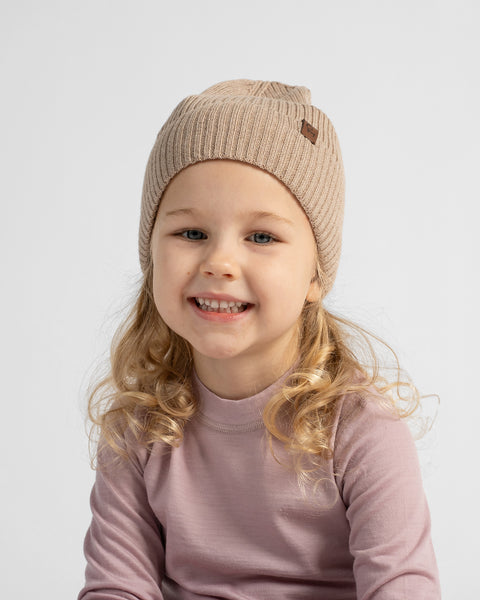 A young child wearing a beige ribbed knit beanie with a small brown logo patch and a dusty rose long-sleeve top, photographed against a light gray studio background.