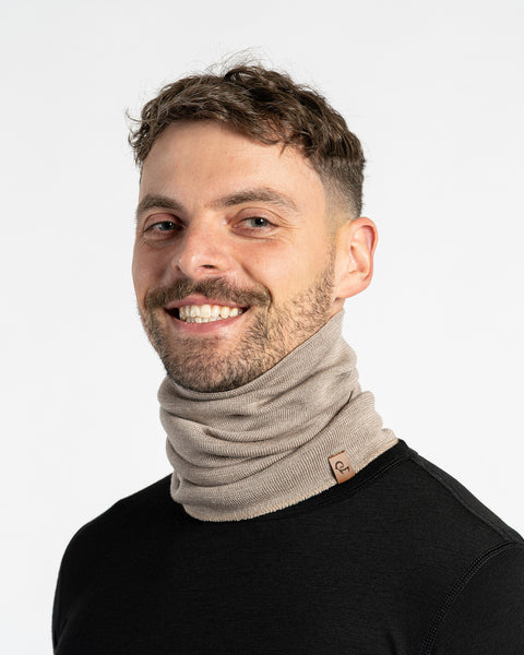 A smiling man with short dark hair and a beard, wearing a creamy beige knit neck warmer and a black long-sleeved shirt, looks directly at the camera. A small brown leather tag is visible on the front of the neck warmer. The background is plain white.