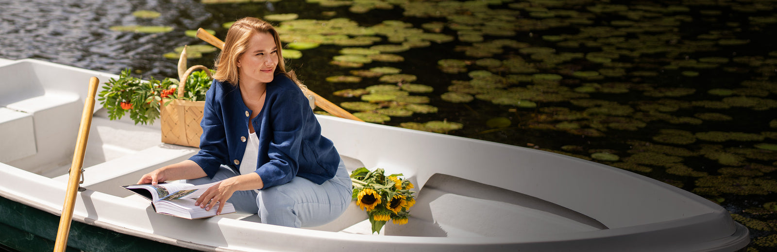 A blonde woman sits in a white rowboat on a lily pad-covered pond, looking thoughtfully off to the right with a gentle smile. She holds an open book in her lap and is wearing a navy blue button-up jacket over a white top with light blue trousers. A wicker basket of greenery and a bouquet of sunflowers rest nearby inside the boat.
