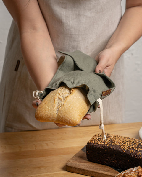 Person holding a loaf of bread wrapped in a reusable bread bag on a wooden table.