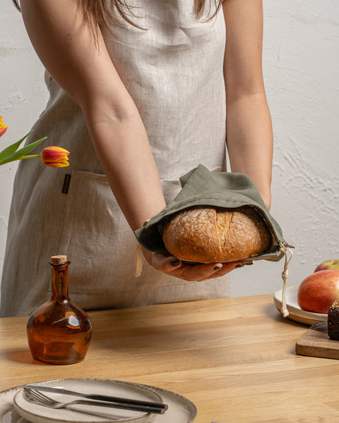 Person holding a loaf of bread wrapped in a green cloth on a wooden table with fruits and a vase.