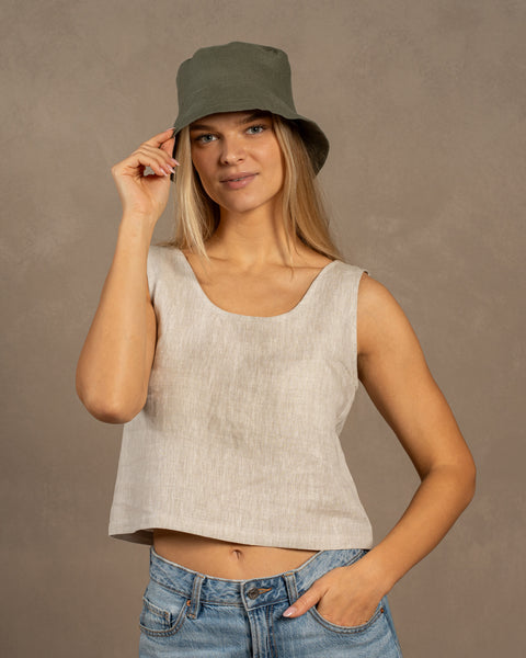 A smiling woman standing confidently with one hand in her blue jeans pocket and the other gently touching the brim of her olive green bucket hat. She is wearing a textured, light beige, sleeveless linen top. The background is a solid, warm grey-brown.
