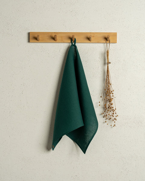 The menique linen kitchen dish towel in the color of dark green hanging on a wooden rack on a wall.
