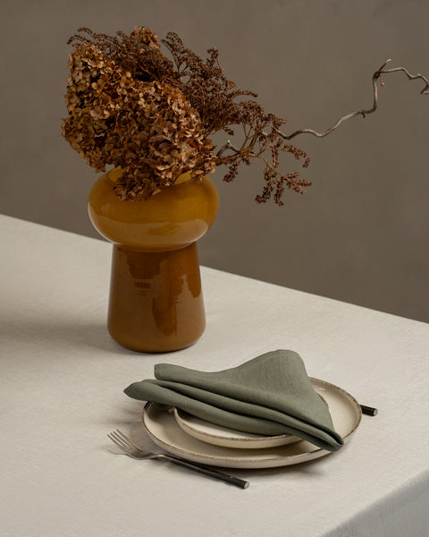 A tabletop covered with a linen tablecloth, with two stacked plates and a vase on top of it. The plates have a stone green linen napkin on top of them, and cutlery by the sides.