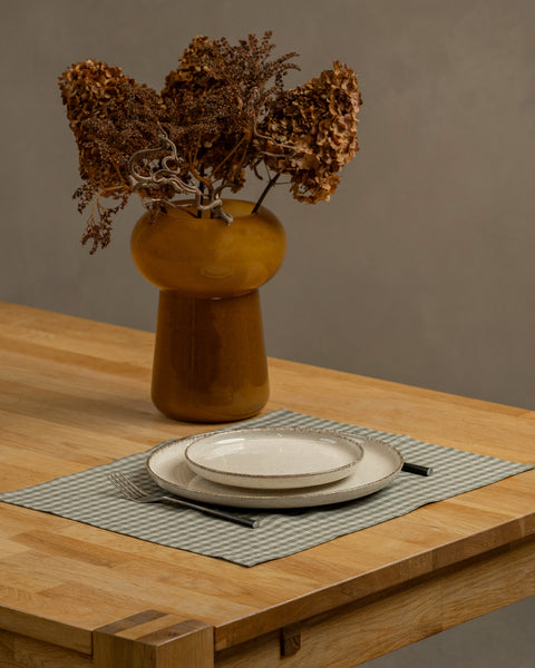 The menique linen placemat in green check color on a wooden table top. There is a vase on top of the table, as well as some cutlery by the sides of the plate, that is on top of the placemat.