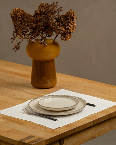 The menique linen placemat in pure white color on a wooden table top. There is a vase on top of the table, as well as some cutlery by the sides of the plate, that is on top of the placemat.