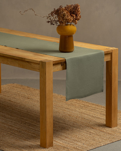 The stone green linen table runner on top of a wooden table, captured in a close up picture from the corner of the table. 