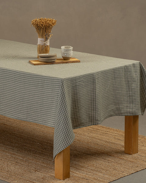 The menique linen tablecloth in green check color, draping over a wooden table, with a vase, wooden board, and a set of dessert plates with tiny clay bowls on top of it.