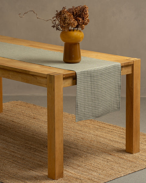 A wooden table positioned on top of a carpet. There is a green check linen table runner and a vase on top.
