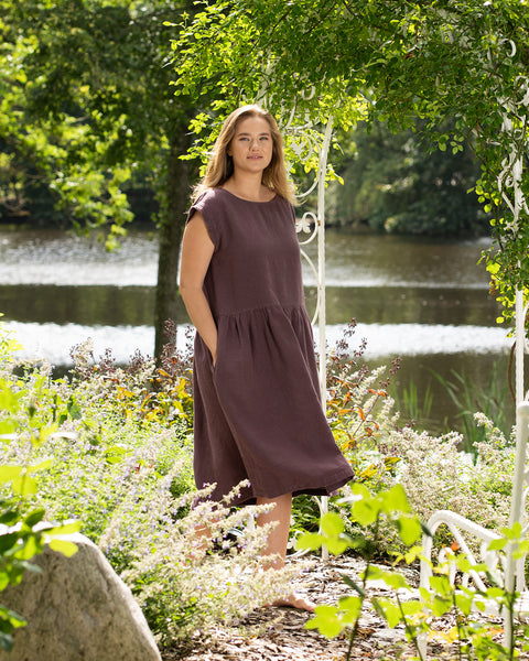 A barefoot woman stands gracefully under a white metal garden archway with a serene lake in the background. She looks directly at the camera, modeling a shadow purple linen dress with a gathered skirt and her hands resting in the side pockets.