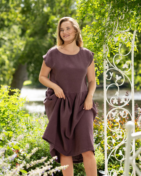 A smiling blonde woman stands in a vibrant garden, gently lifting the sides of her flowing shadow purple linen dress. The breeze catches her hair as she poses naturally near a decorative white archway.