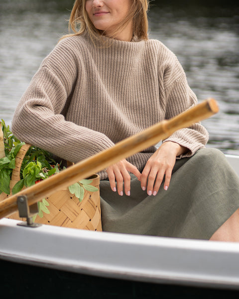 A close-up view shows a woman sitting in a rowboat, dressed in a cozy beige cashmere sweater and a stone green skirt. Her hands rest gently on the skirt near a wooden oar and a basket of fresh greens, with the textured ripples of the lake serving as the background.