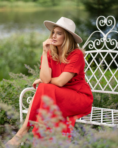 A woman sits thoughtfully on a decorative white garden bench overlooking a lake, resting her hand near her chin as she gazes off to the side. She is elegantly styled in a bright red linen wrap dress and a wide-brimmed light beige hat.
