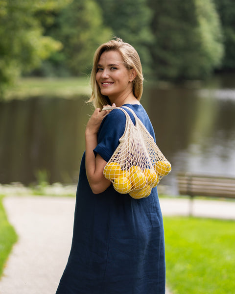A blonde woman smiles warmly over her shoulder while strolling through a lush green park near a lake. She is dressed in a storm blue linen dress and carries a string mesh bag loaded with fresh lemons slung over her back.