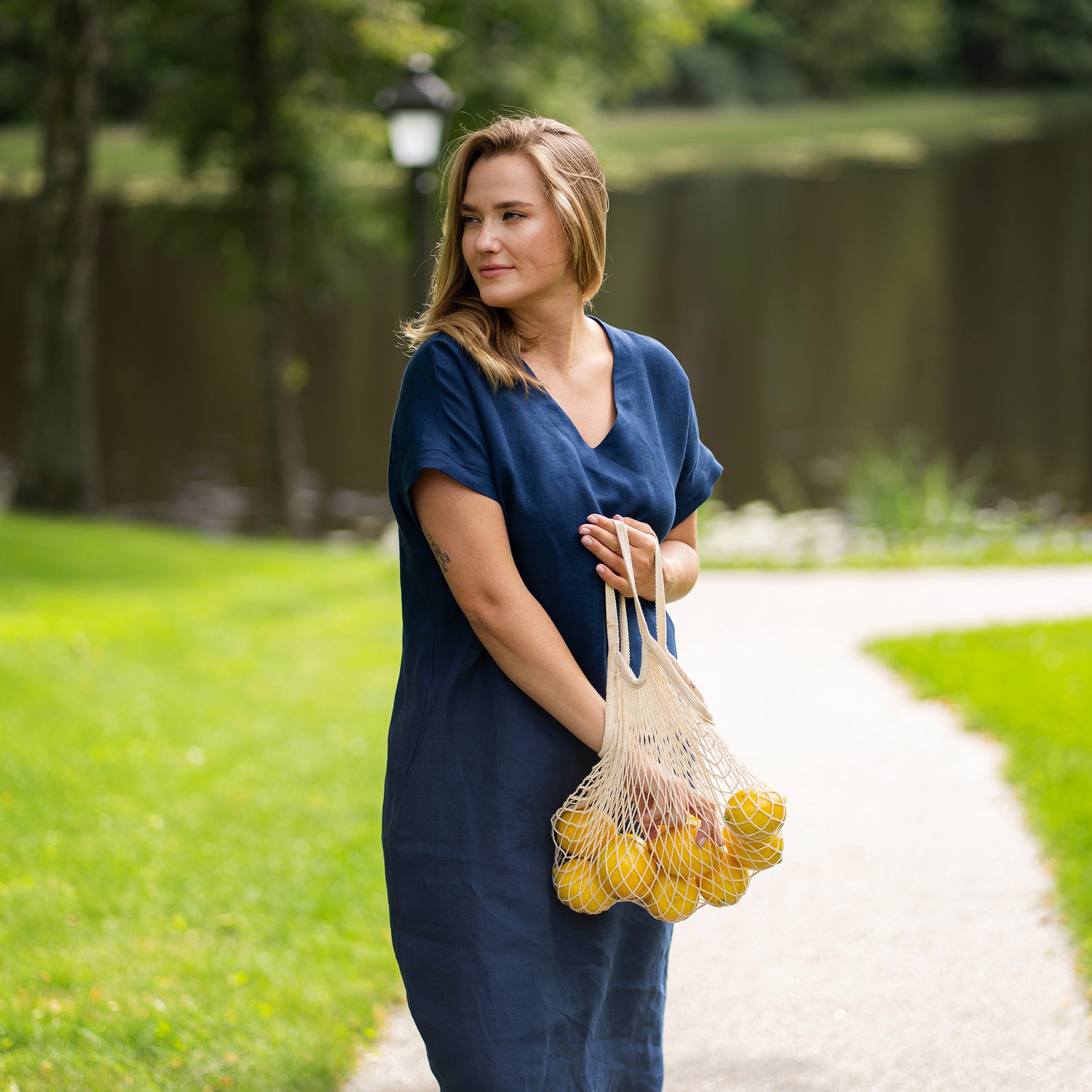 A woman stands on a park pathway holding a string bag of lemons down by her side, looking softly toward the camera. She models a storm blue linen dress with a v-neckline and a tied waist, paired with brown slip-on sandals.