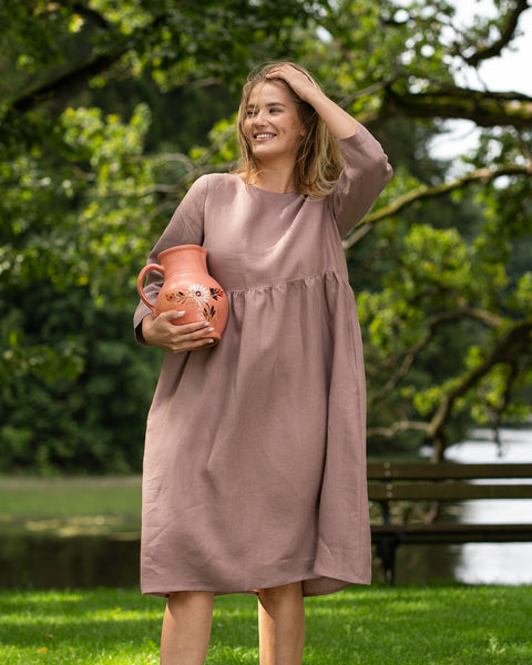 A brightly smiling woman stands outdoors in a scenic park, wearing a faded rose, long-sleeve linen dress. She cradles a terracotta pitcher under one arm and gently brushes a hand through her hair while looking off to the side.