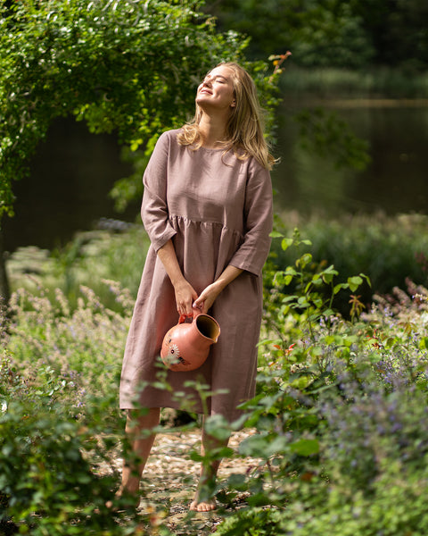 A barefoot woman stands in a lush, sunlit garden wearing a long-sleeved, faded rose linen dress with a gathered waist. She holds a painted terracotta pitcher in front of her, tilting her head back with closed eyes to soak in the sunshine.