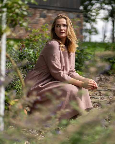 A woman wearing a faded rose linen dress crouches on a stone pathway surrounded by lush garden greenery. She rests her clasped hands gently on her knees while looking thoughtfully off to the side.
