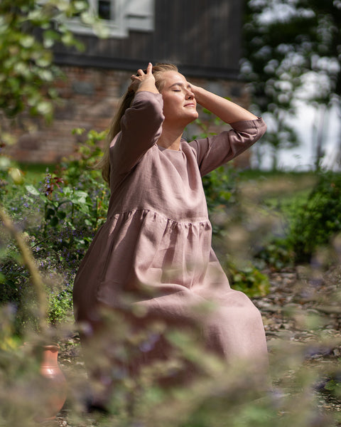 A woman wearing a faded rose linen dress kneels among blooming garden plants with her face turned upwards toward the sun. She rests both hands in her hair, enjoying a serene moment outdoors with her eyes gently closed.