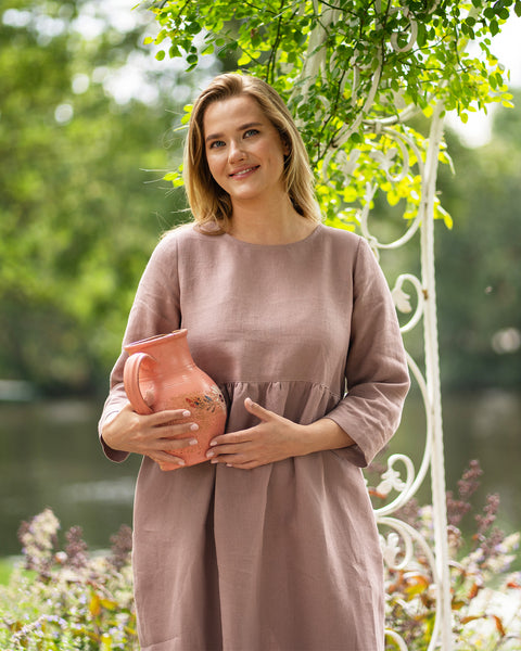 A woman stands gracefully in a green, tree-filled park wearing a long-sleeve, faded rose linen dress. She holds a terracotta pitcher against her waist with both hands, gazing thoughtfully off to the side.