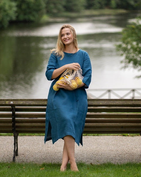 A barefoot woman sits comfortably on a wooden park bench beside a tranquil lake, smiling as she looks off to the side. She wears a cobalt blue linen dress with side slits and rests a string bag filled with bright yellow lemons softly on her lap.
