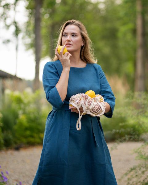 A woman stands on a park pathway, thoughtfully holding a bright yellow lemon near her chin as she looks into the distance. She is dressed in a cobalt blue linen dress with a tied waist, gently cradling a string bag of lemons in her arms.