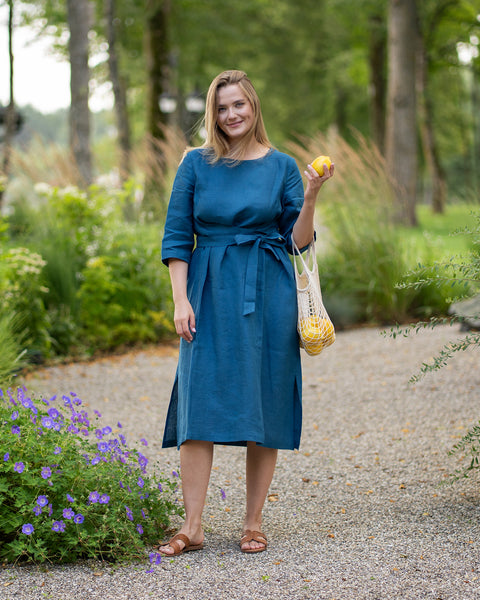 A smiling woman stands on a gravel pathway, wearing a vibrant cobalt blue linen dress with a waist tie and side slits. She looks directly at the camera, casually holding a fresh lemon and a string bag full of lemons in her left hand.