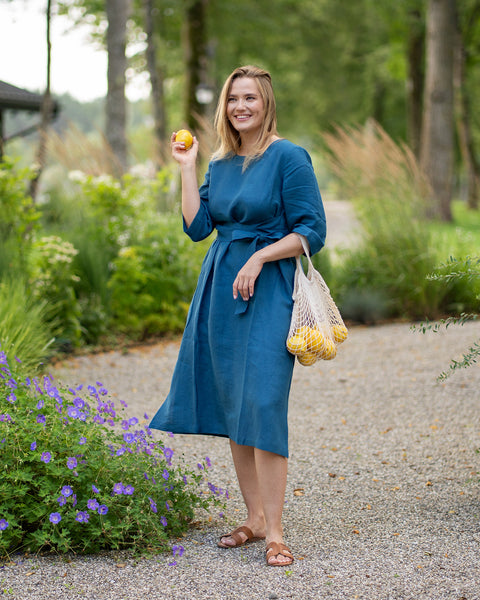 A smiling woman stands on a gravel garden path wearing a cobalt blue, three-quarter sleeve linen dress with a tied waist. She playfully holds up a single lemon in one hand while carrying a string bag full of lemons in the other.