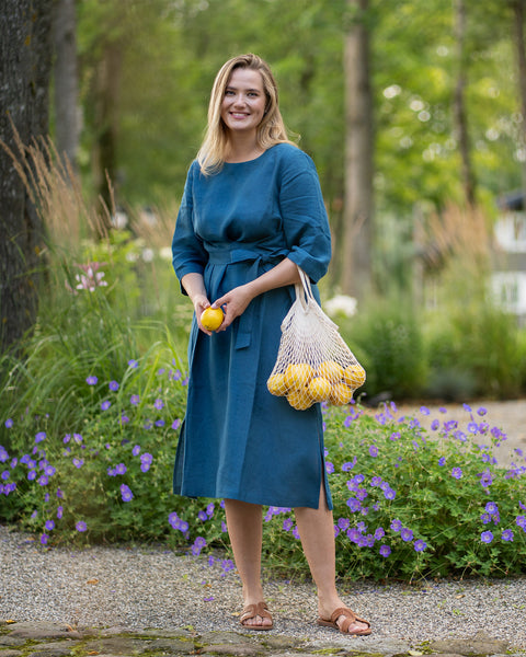 A smiling woman stands on a gravel pathway surrounded by vibrant garden flowers and greenery. She models a cobalt blue linen midi dress with a tied waist, holding a lemon in one hand and a string mesh bag of lemons in the other.