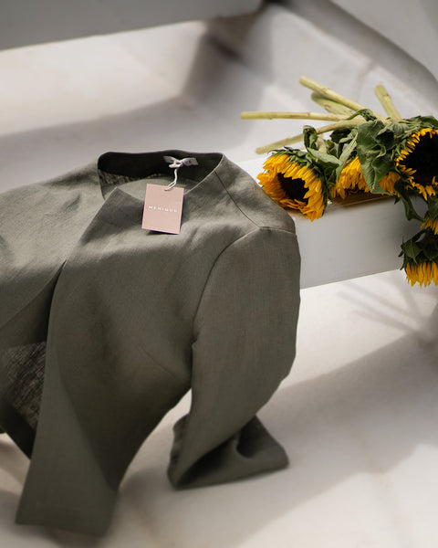 A neatly folded stone green linen jacket rests across a white surface in the sunlight, displaying a soft pink brand tag. A cluster of bright yellow sunflowers lies just behind the collar, beautifully contrasting with the earthy green fabric.