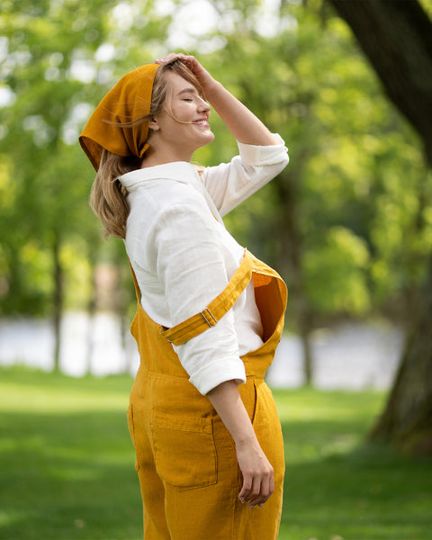 A smiling woman stands in a lush green park, turning her face toward the sun with her eyes closed while gently touching her head. She is wearing a pure white linen shirt layered under a vibrant, spicy yellow linen jumpsuit, accessorized with a matching yellow bandana.