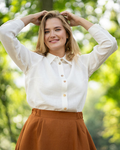 A blonde woman beams at the camera while standing outdoors against a backdrop of lush green trees. She rests both hands gracefully on top of her head while wearing a white shirt and brown skirt.