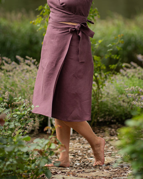 A woman stands barefoot on a garden pathway, gently lifting one heel. Her outfit features a shadow purple linen wrap skirt and a matching crop top with a waist tie, blending beautifully into the surrounding greenery.