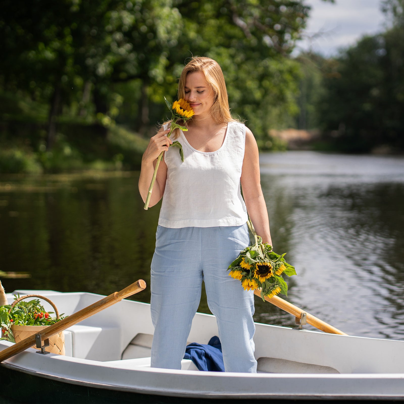 A woman reclines gracefully inside a small white boat, closing her eyes and soaking up the sunlight with one hand gently resting in her hair. She is dressed comfortably in a white sleeveless linen top and light blue pants, sitting right beside a filled harvest basket.
