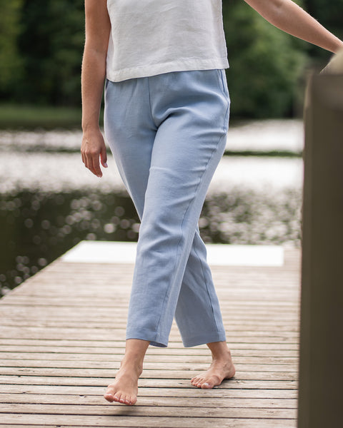 A woman is shown from the chest down wearing a white linen top and light blue linen trousers. She poses barefoot with her legs slightly crossed on a wooden dock beside a calm lake.