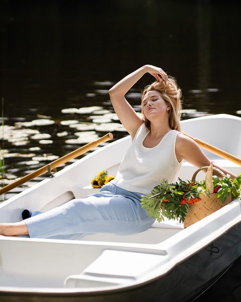 A woman lounges comfortably in a white rowboat, resting her hand behind her head. A basket full of fresh greens sits on the seat beside her as she relaxes on the water.