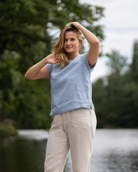 A woman stands on a wooden dock near a river, running her hands through her hair while smiling warmly at the camera. She is dressed comfortably in a loose-fitting, light blue short-sleeved top and beige pants.