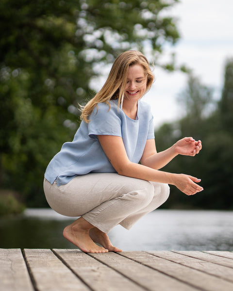A woman crouches barefoot on a wooden dock by a calm body of water, smiling cheerfully with her hands gently held in front of her. She is wearing a light blue short-sleeved top and natural beige trousers.