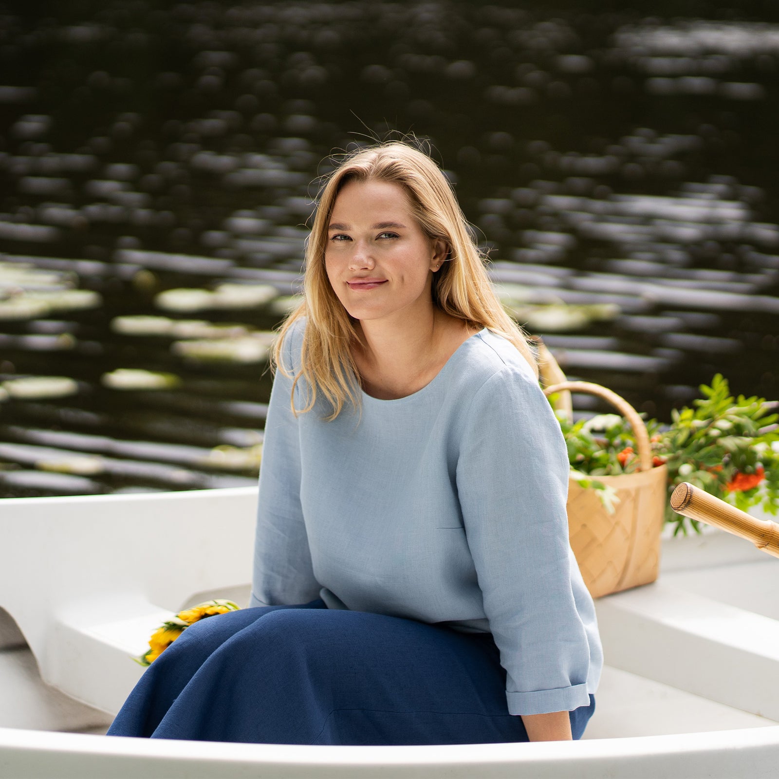 A woman sits in a white boat with her arms stretched joyfully up towards the sky, her eyes closed in relaxation. She is wearing a light blue linen shirt and dark blue pants, accompanied by bright sunflowers on the seat next to her.