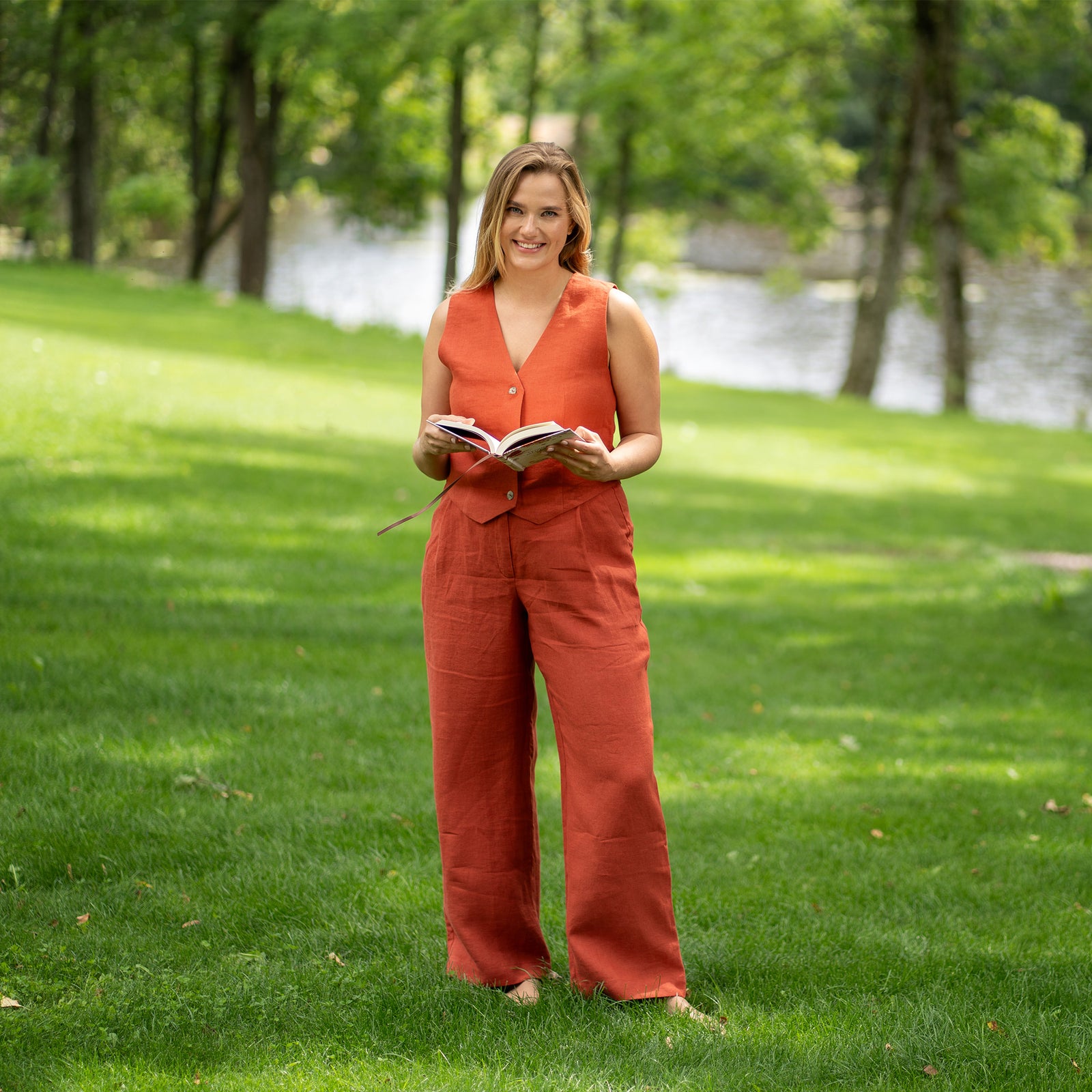 A woman in a stylish cinnamon red linen vest and trousers stands outdoors holding a book open in her hands. She looks towards the camera with a subtle smile, set against a vibrant green, soft-focus background of park trees.