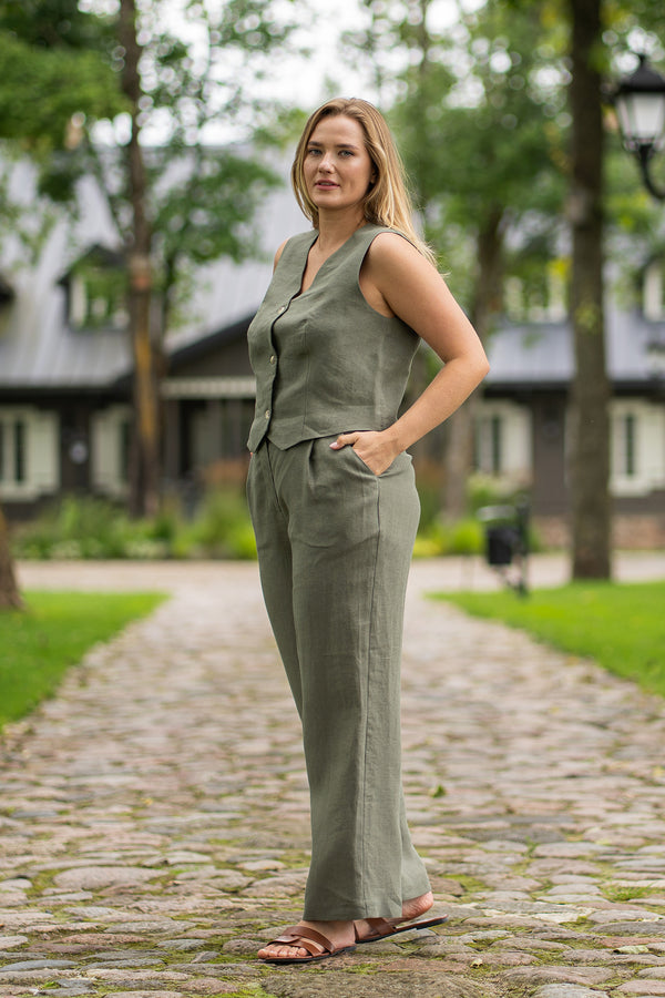 Standing outdoors on a scenic stone path, a woman poses with both hands tucked into the pockets of her stone green linen pants. She wears a matching sleeveless button-up vest and looks warmly toward the camera.