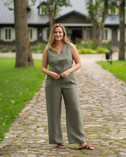 A woman stands confidently on a cobblestone walkway in a park-like setting with a traditional building in the background. She is dressed in a matching stone green outfit featuring a linen vest and wide-leg trousers, smiling softly with her hands resting near her waist.