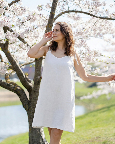 Woman wearing pure white linen slip dress with tie shoulder straps, lightweight breathable summer dress styled outdoors under blooming trees