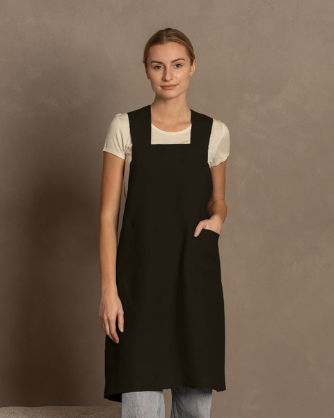 A woman with fair skin and blonde hair pulled back looks directly at the camera. She is wearing a white short-sleeved shirt under a pure black linen cross-back apron with a front pocket. Light blue jeans are visible below the apron. She stands against a plain warm brown background.