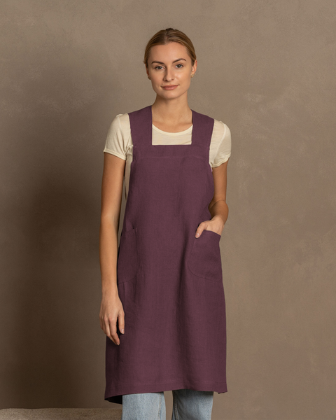 A fair-skinned woman with blonde hair pulled back looks directly at the viewer. She is wearing a white short-sleeved shirt under a shadow purple linen cross-back apron with front pockets. Light blue jeans are visible below the apron. She stand against a plain warm brown background.